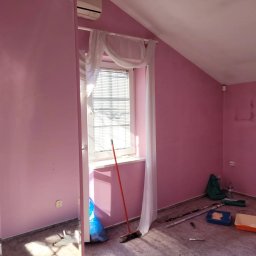 Usługi Malarskie Warszawa Robert Gawlik - Różowy pokój po odświeżeniu, widoczne narzędzia malarskie, zasłonięte okno z żaluzjami, ślady po poprzednim malowaniu na ścianie