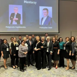 Grupa osób pozuje do zdjęcia na tle ekranu z wizerunkami mentorów, Małgorzaty Cegiełkowskiej i Przemysława Ciołaka, podczas wydarzenia networkingowego.