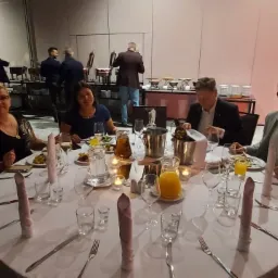 Bankiet w sali konferencyjnej: Uczestnicy siedzą przy okrągłym stole z nakryciem, na którym stoją szklanki, talerze z jedzeniem i napoje. W tle bufet i obsługa.