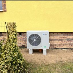 JCC Robert Kamiński - Zewnętrzna jednostka klimatyzacyjna York na betonowych podstawach, umieszczona przy żółtej ścianie budynku z ceglaną podmurówką, widoczna z boku krzewu.