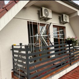 JCC Robert Kamiński - Antena telewizyjna zamontowana na balkonie domu jednorodzinnego, widoczne jednostki zewnętrzne klimatyzacji na ścianie, balkon z kwiatami.