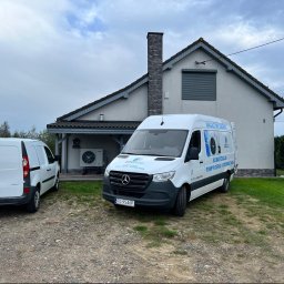 JCC Robert Kamiński - Dwa białe busy zaparkowane przed domem jednorodzinnym; na jednym busie widoczne napisy: Magazyny energii, Klimatyzacja, pompy ciepła, fotowoltaika, oraz logo firmy JCC.