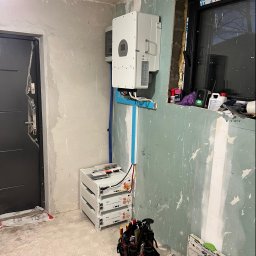 JCC Robert Kamiński - Wnętrze w trakcie remontu z zamontowanym inwerterem na ścianie, trzema modułami bateryjnymi Deye ustawionymi na podłodze oraz torbą narzędziową obok, widoczne przewody i niebieska rura instalacyjna.