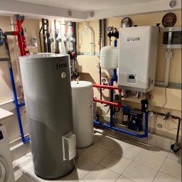 JCC Robert Kamiński - Kotłownia z szarym i białym zasobnikiem ciepłej wody marki Ferroli i York, orurowanie w kolorze niebieskim i czerwonym, na ścianie filtry i liczniki, płytki na podłodze.