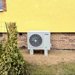 JCC Robert Kamiński - Biała jednostka zewnętrzna pompy ciepła marki York, umieszczona na betonowych podstawach przy żółtej ścianie domu z ceglaną podmurówką, widoczna instalacja rurowa i krzew z ozdobnymi liśćmi.