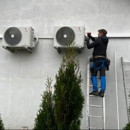 JCC Robert Kamiński - Montaż klimatyzacji Rotenso na jasnej ścianie budynku, instalator na drabinie z wiertarką, widoczne dwie jednostki zewnętrzne i tuje w tle.