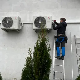 Montaż klimatyzacji Rotenso na jasnej ścianie budynku, instalator na drabinie z wiertarką, widoczne dwie jednostki zewnętrzne i tuje w tle.