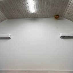 JCC Robert Kamiński - Dwa białe klimatyzatory zamontowane symetrycznie na białej ścianie pod drewnianym sufitem. Po prawej stronie, pod sufitem, wisi złoty balon.