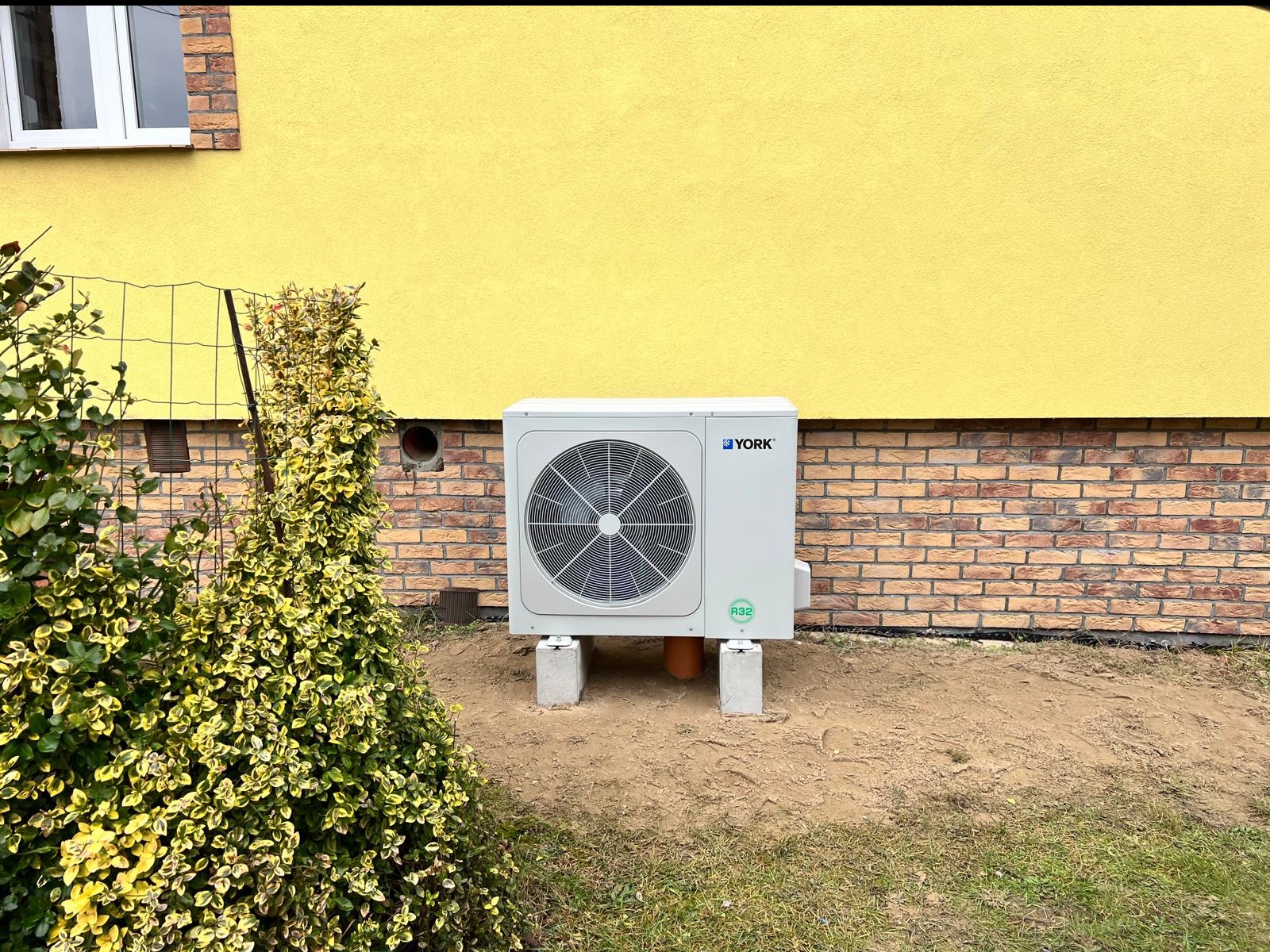 Zewnętrzna jednostka klimatyzacyjna York na betonowych podstawach, umieszczona przy żółtej ścianie budynku z ceglaną podmurówką, widoczna z boku krzewu.