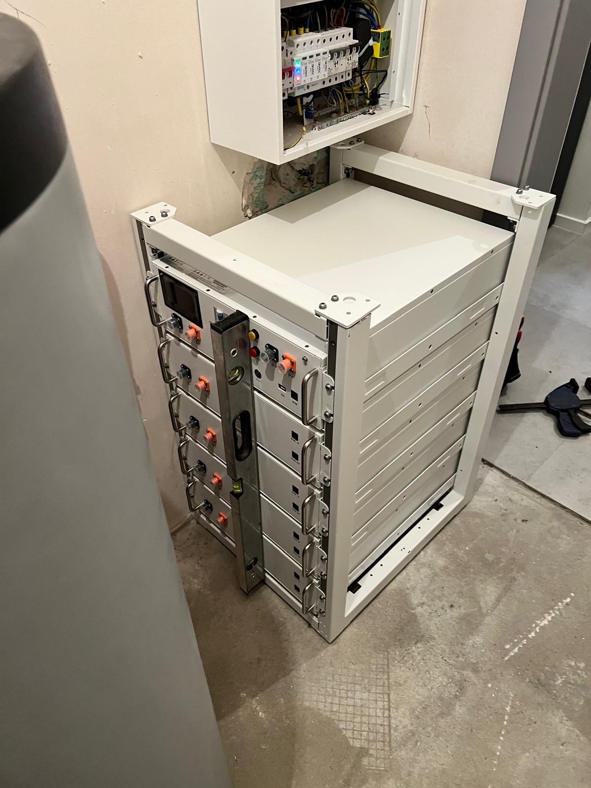 Biały, modułowy magazyn energii ustawiony przy ścianie, poziomica oparta o jego bok, skrzynka elektryczna nad nim.