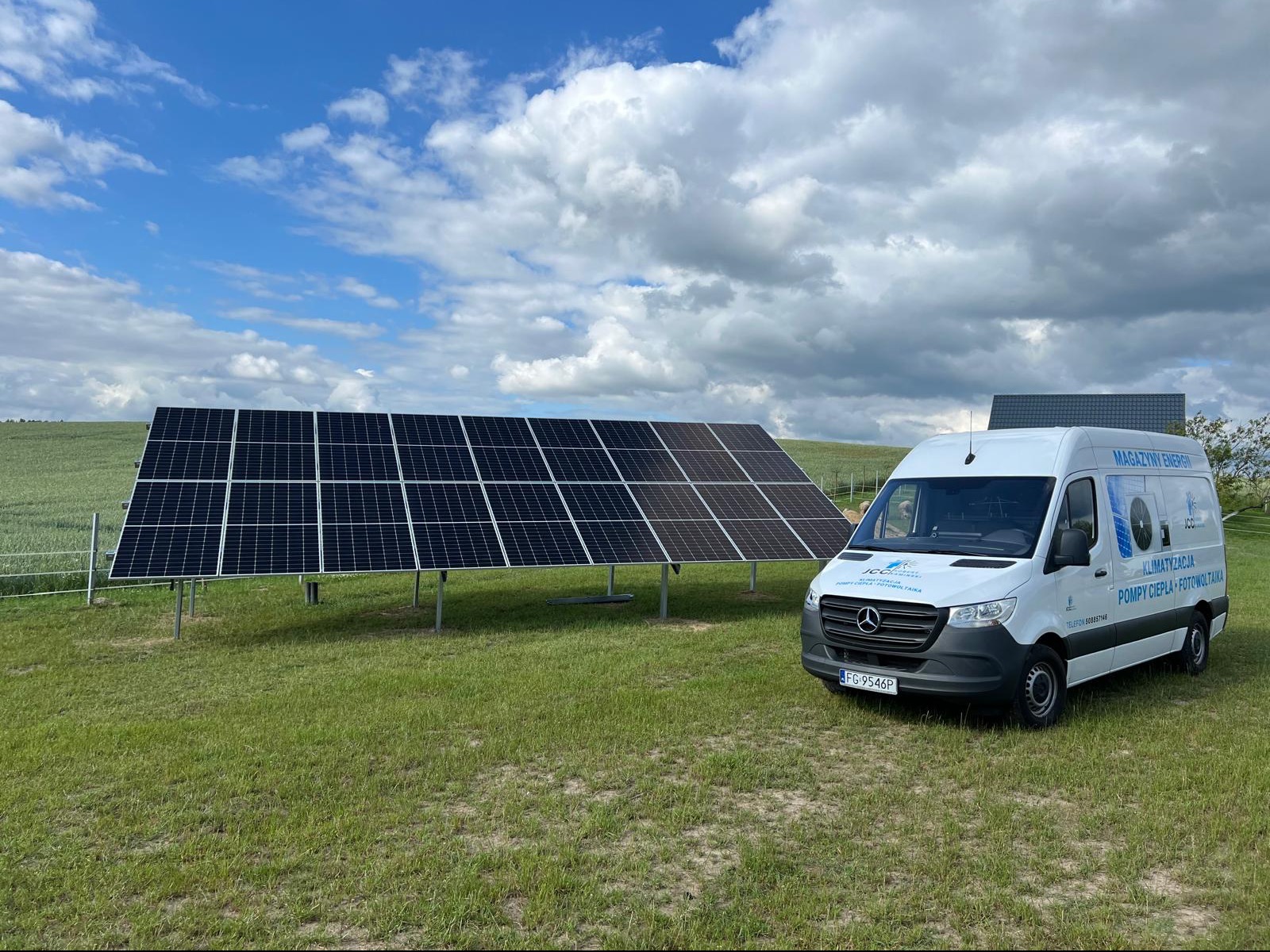Instalacja fotowoltaiczna na łące z widocznym białym busem firmy oferującej magazyny energii, klimatyzację, pompy ciepła i fotowoltaikę. Tło stanowi błękitne niebo z obłokami.