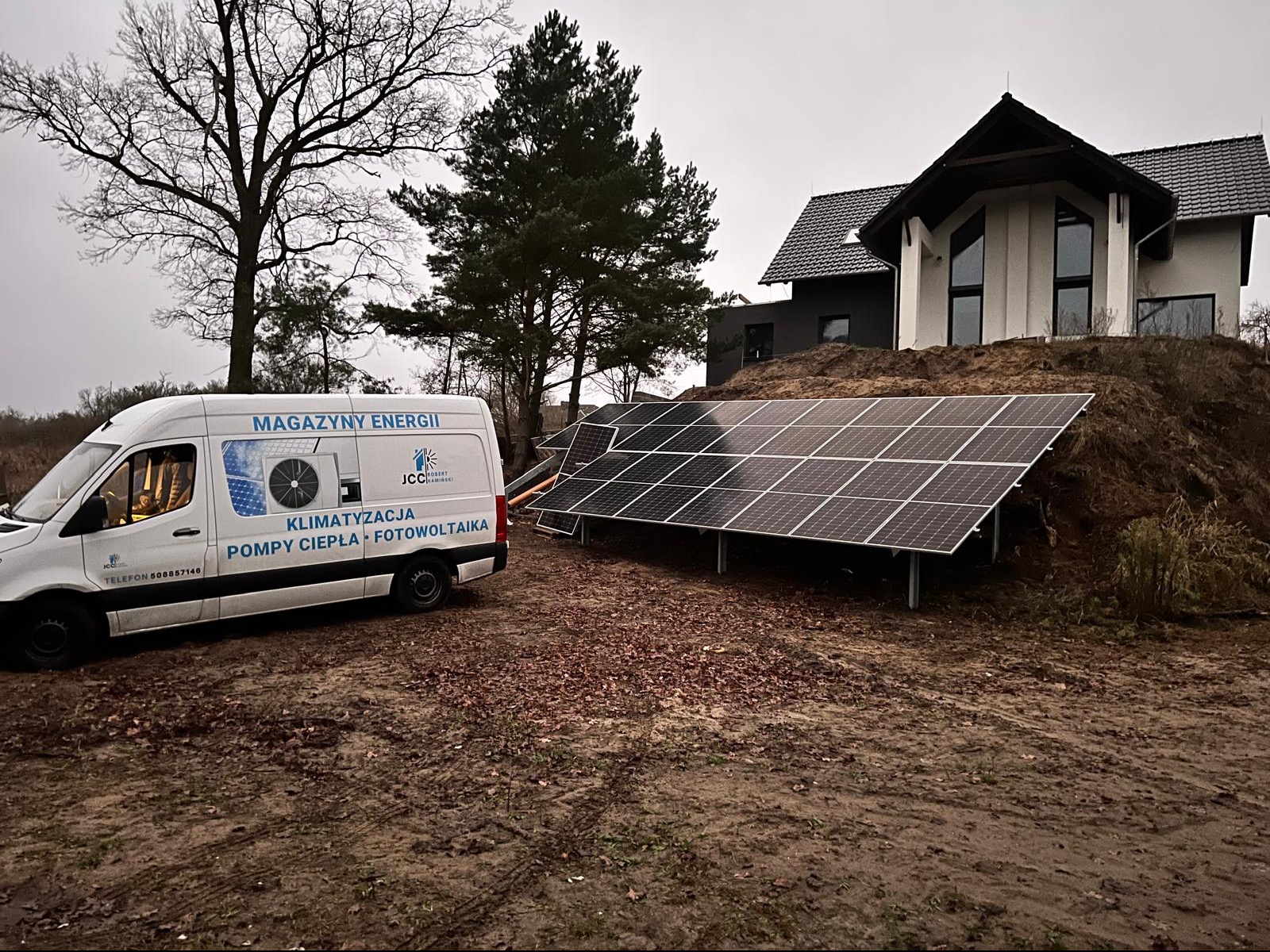 Instalacja paneli fotowoltaicznych na wzgórzu obok nowoczesnego domu, z widocznym samochodem dostawczym firmy oferującej magazyny energii, klimatyzację, pompy ciepła i fotowoltaikę.