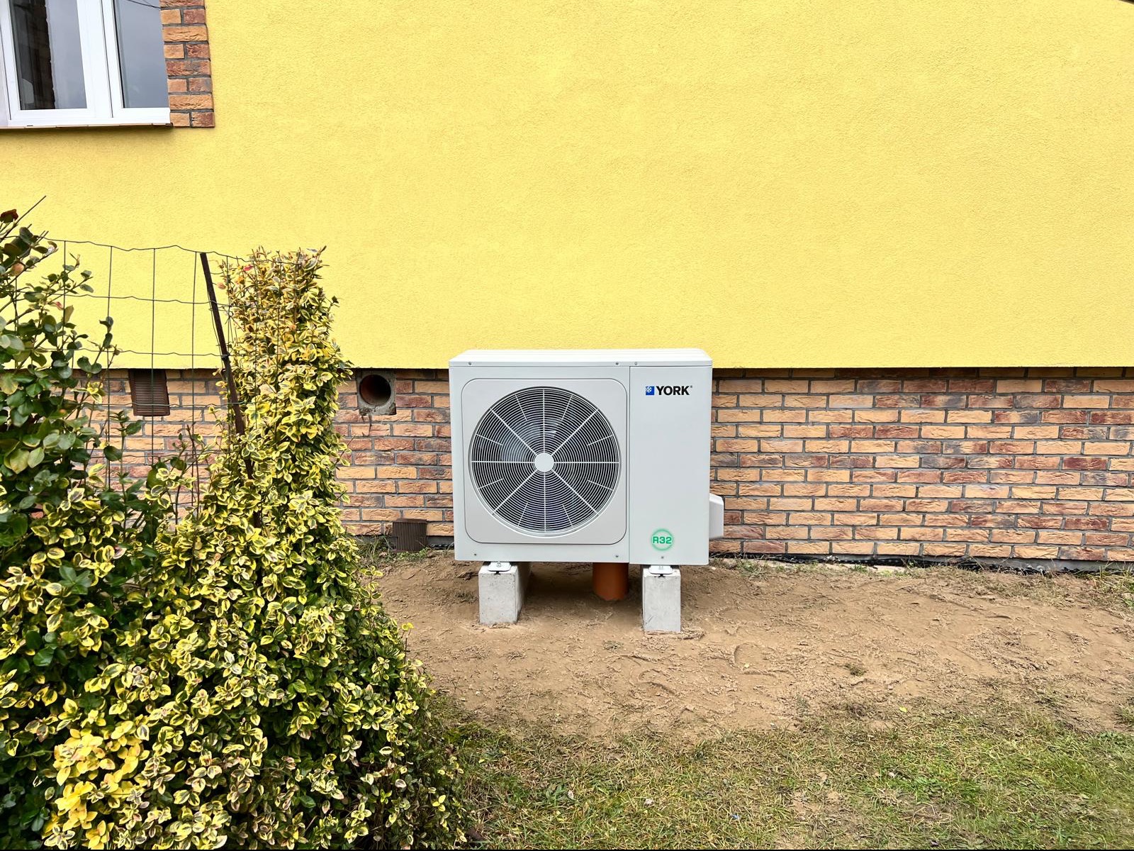 Biała jednostka zewnętrzna pompy ciepła marki York, umieszczona na betonowych podstawach przy żółtej ścianie domu z ceglaną podmurówką, widoczna instalacja rurowa i krzew z ozdobnymi liśćmi.