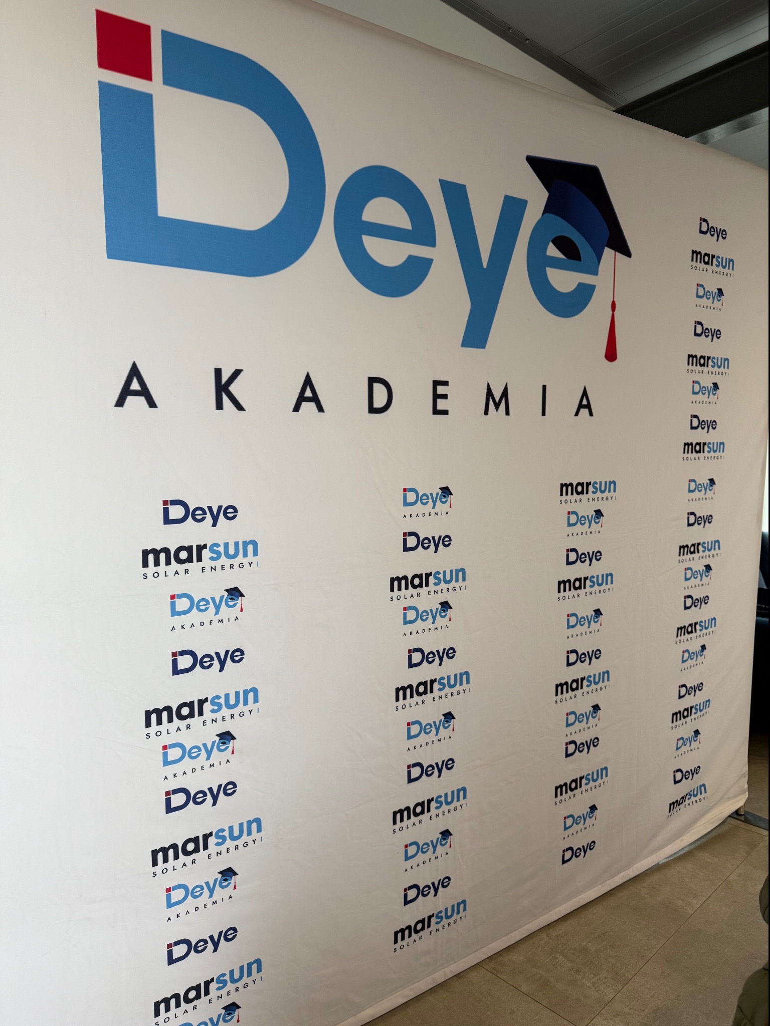 Tło reklamowe z powtarzającym się logo firmy Deye i Marsun Solar Energy oraz logotypem Deye Akademia z motywem czapki absolwenta.