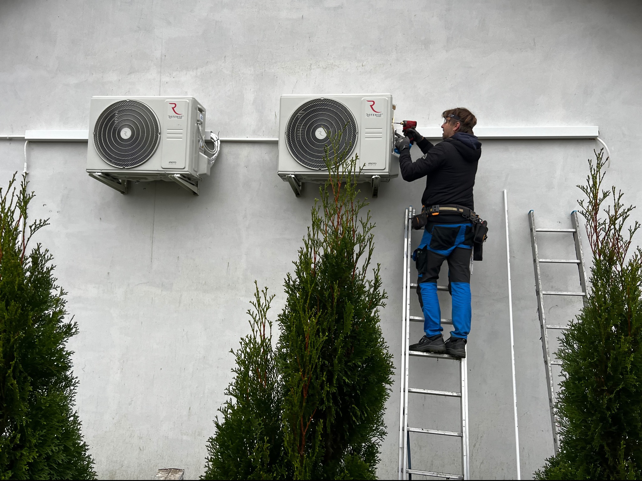 Montaż klimatyzacji Rotenso na jasnej ścianie budynku, instalator na drabinie z wiertarką, widoczne dwie jednostki zewnętrzne i tuje w tle.
