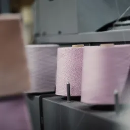 Rzędy szpul z delikatną, różową przędzą na nowoczesnej maszynie dziewiarskiej, z ostrością skupioną na środkowej szpuli i subtelnym rozmyciem tła.