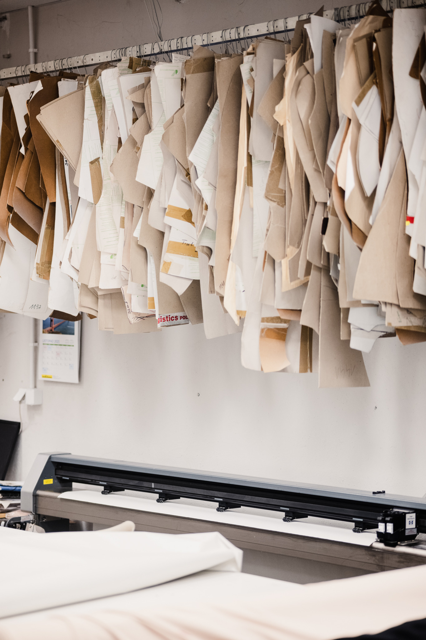 Pracownia krawiecka: wiszące papierowe wykroje odzieży nad ploterem tnącym materiał, kalendarz na ścianie w tle.