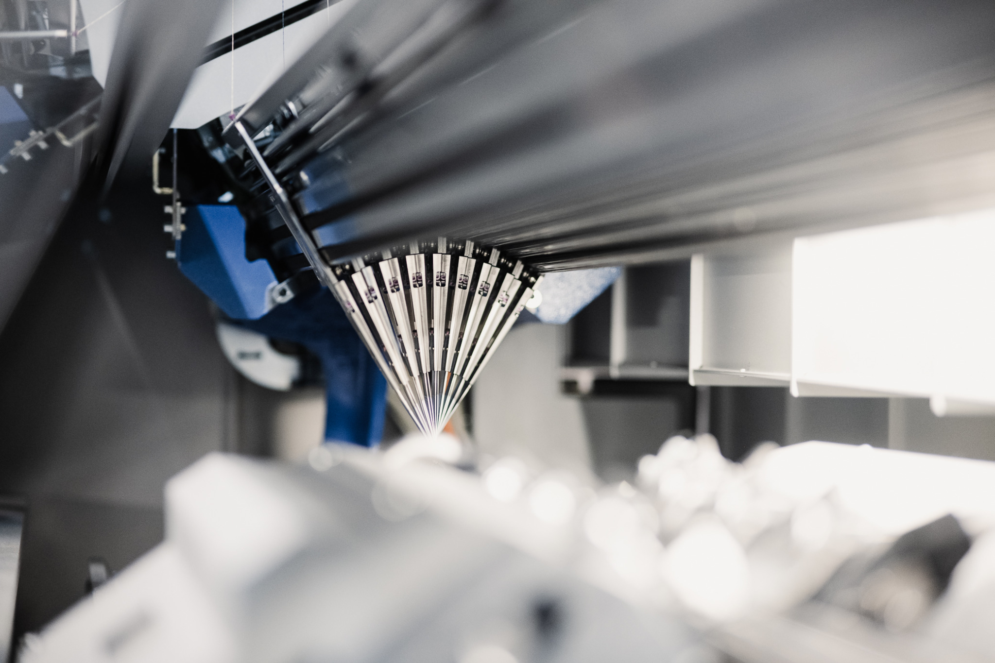 Wnętrze przemysłowej maszyny dziewiarskiej z widocznymi igłami i prowadnicami nici, ujęcie z dołu w jasnym oświetleniu.