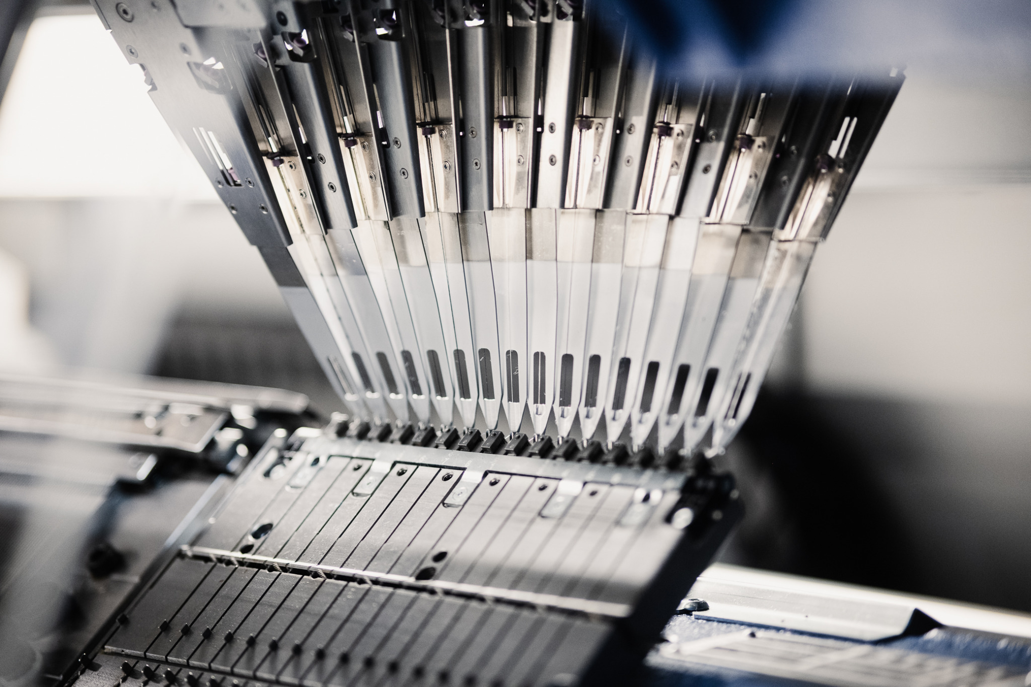 Metalowe elementy maszyny dziewiarskiej w zbliżeniu, widoczne igły i prowadnice, oświetlone z góry, z rozmytym tłem.