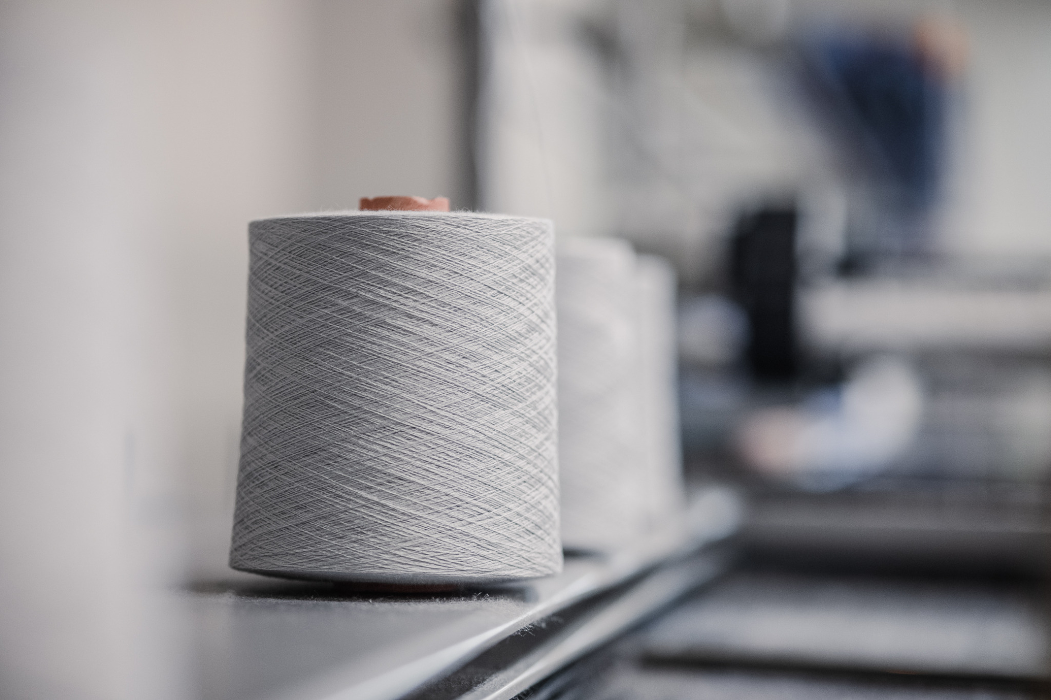 Szpula szarej nici na metalowej półce, kolejne szpule rozmyte w tle, widoczne ślady użytkowania na półce.