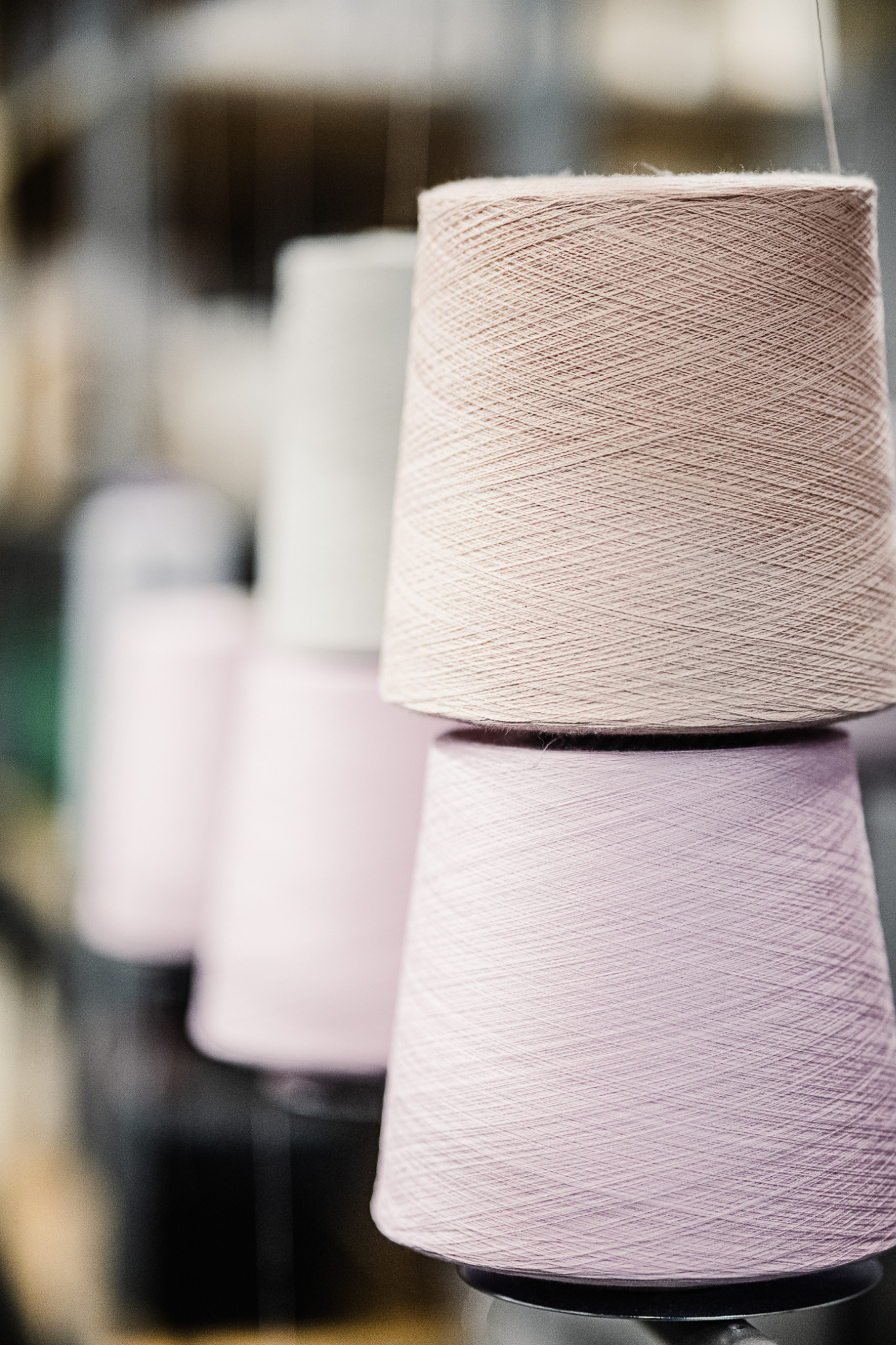 Ustawione jeden na drugim stożkowe szpule z nici w odcieniach beżu i pudrowego różu, z delikatnym światłem padającym na fakturę przędzy.