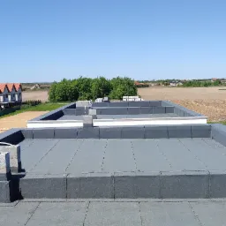 Widok z góry na płaski dach pokryty szarą papą, z widocznymi obróbkami blacharskimi i elementami wentylacyjnymi na tle błękitnego nieba i zabudowań w oddali.