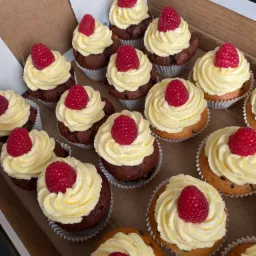 Kartonowe pudełko pełne babeczek z kremem waniliowym i malinami, część babeczek ma ciemne, czekoladowe ciasto, a część jasne z kawałkami czekolady, widoczne papierowe foremki.