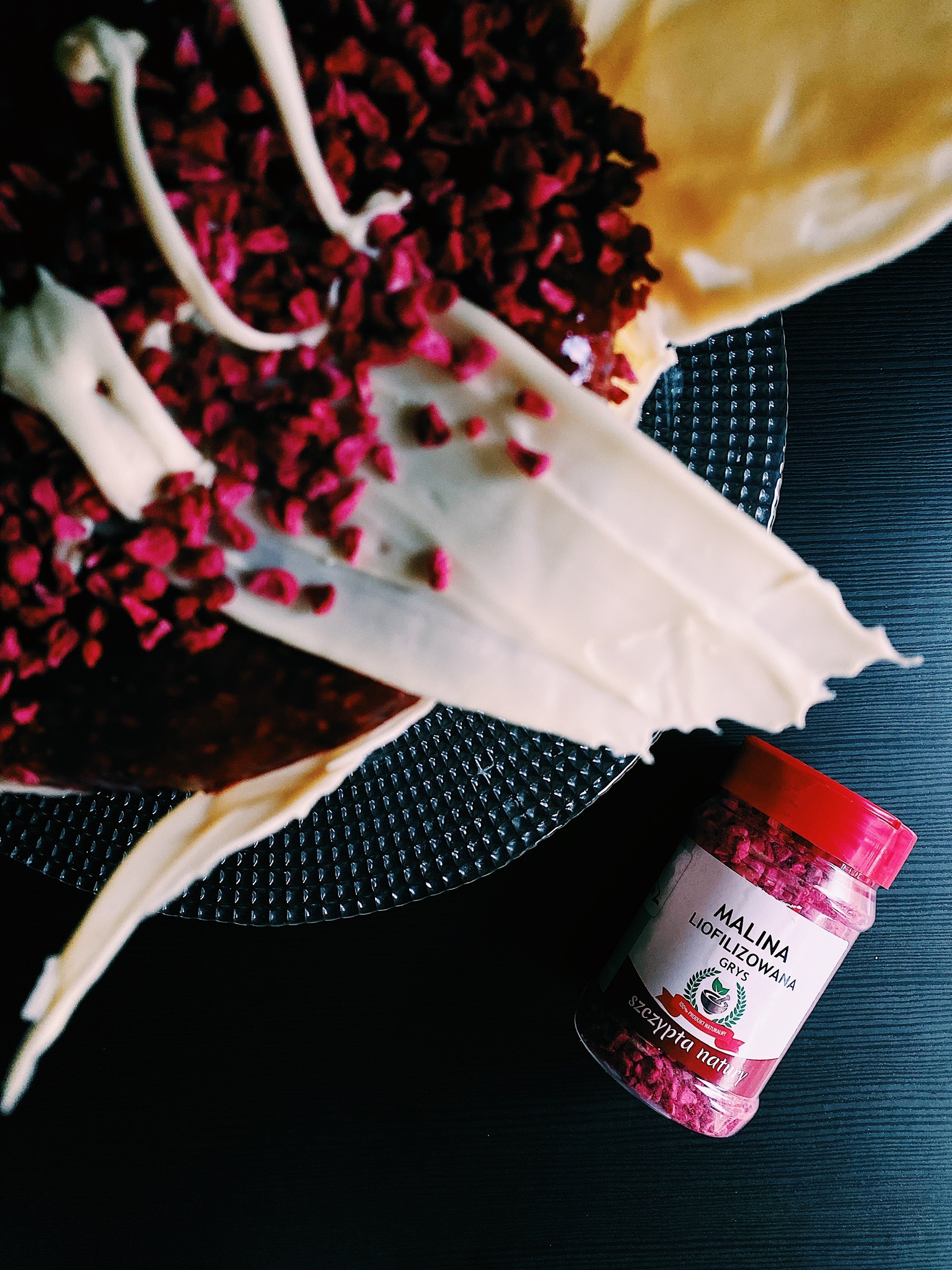 Dekorowany tort z polewą z białej czekolady i liofilizowanymi malinami na szklanym talerzu obok słoiczka z liofilizowanymi malinami w grysie.