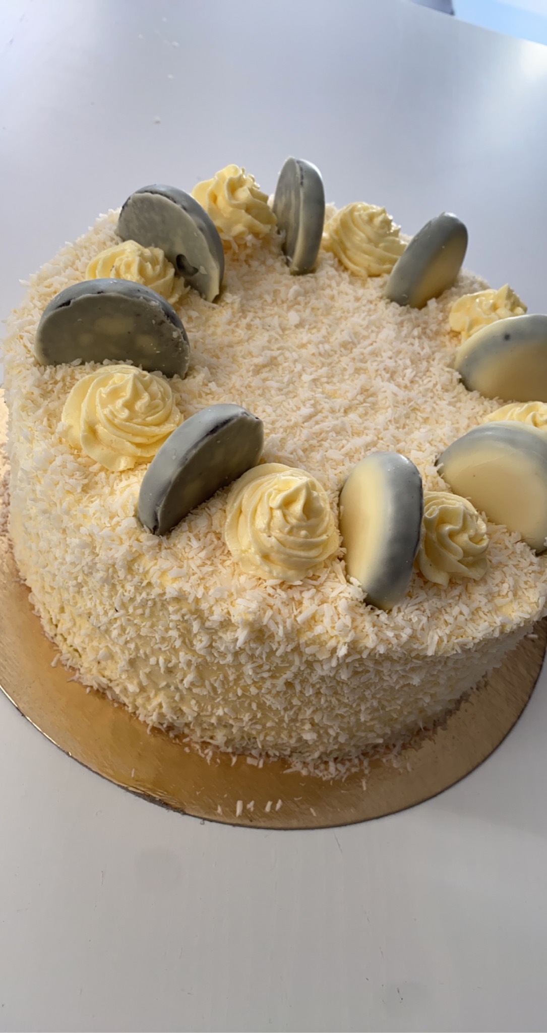 Okrągły tort kokosowy z kremowymi rozetkami i dekoracjami z białej i szarej czekolady, umieszczony na złotej podkładce.