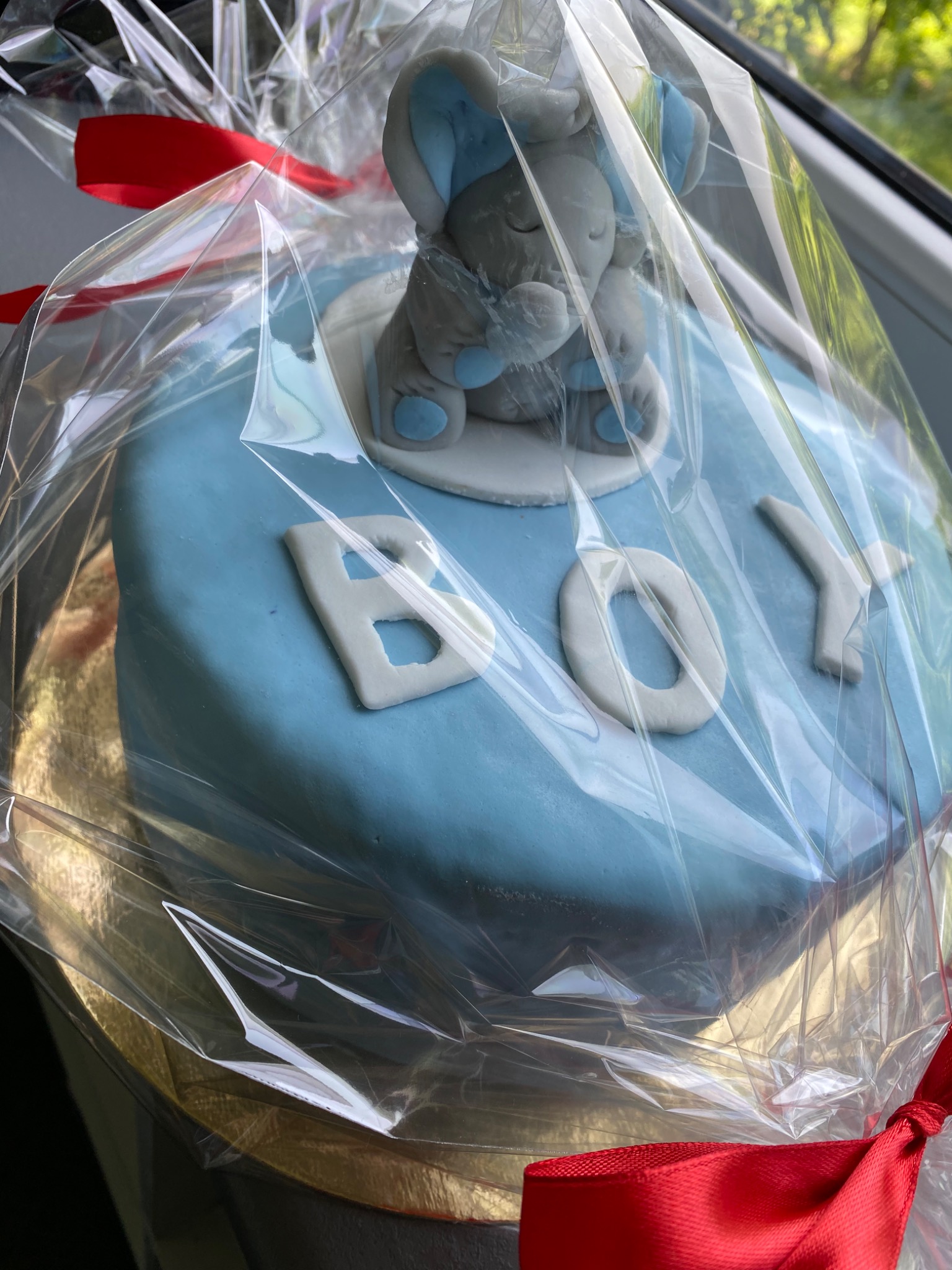 Jasnoniebieski tort z napisem 'BOY' i figurką śpiącego słonia, zapakowany w przezroczysty celofan z czerwoną kokardą.