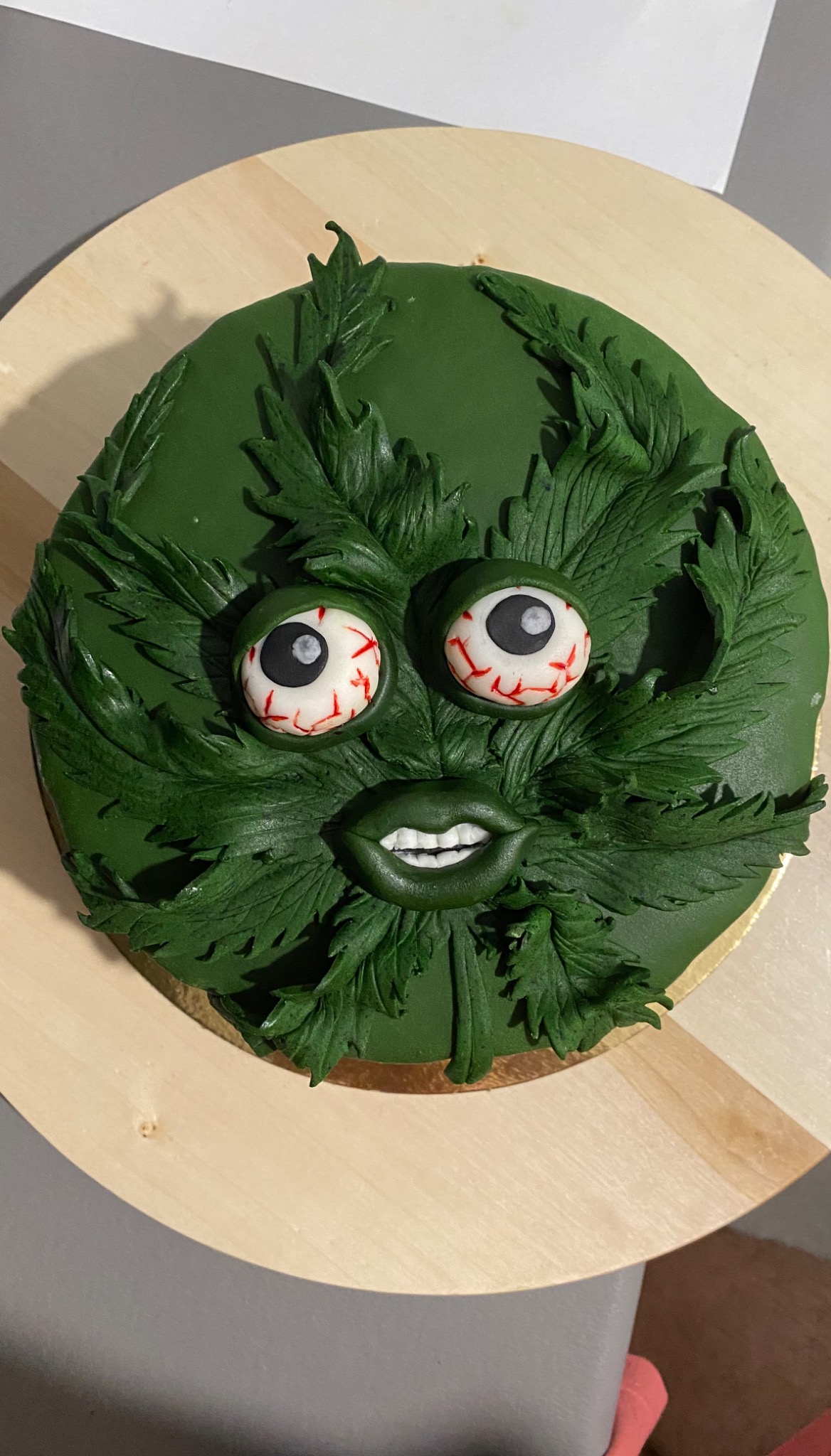 Okrągły tort z zielonego lukru, udekorowany liśćmi marihuany z lukru, oczami z czerwonymi żyłkami i ustami z białymi zębami, całość umieszczona na drewnianej podkładce.