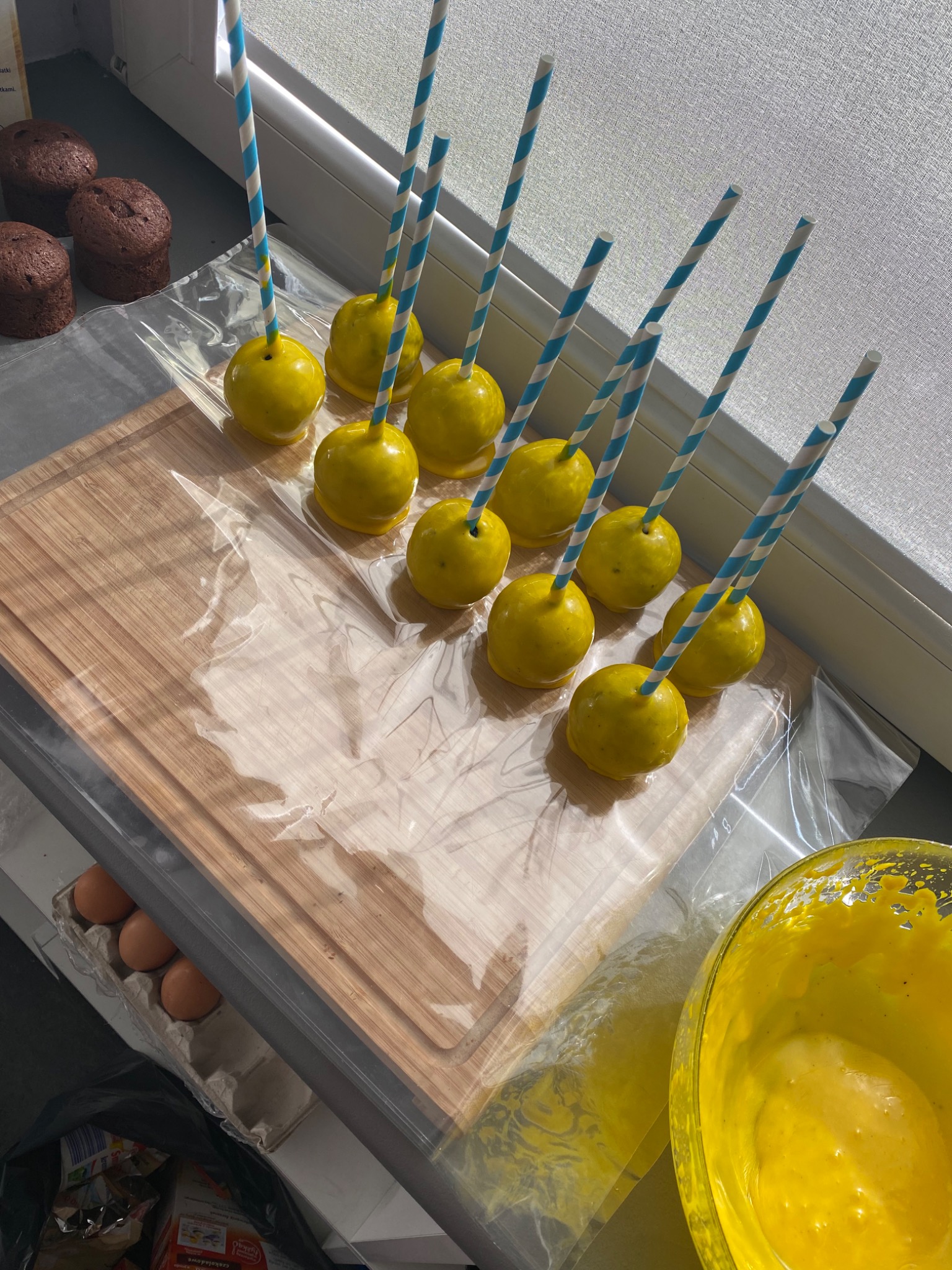 Przygotowanie lizaków cake pops: żółte kulki na patyku, ułożone na desce do krojenia wyłożonej folią, obok miska z żółtą polewą i foremki do babeczek.
