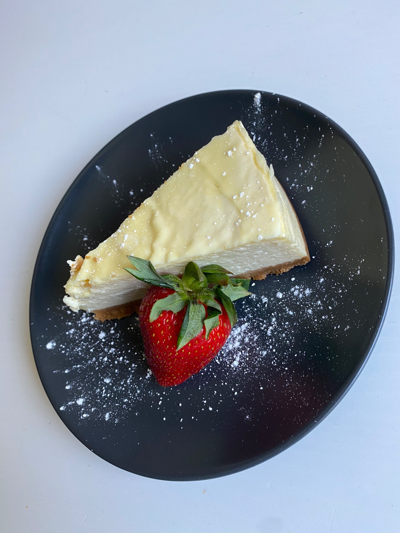 Kremowy sernik nowojorski w trójkącie, posypany cukrem pudrem, podany na czarnym talerzu z dużą, świeżą truskawką.