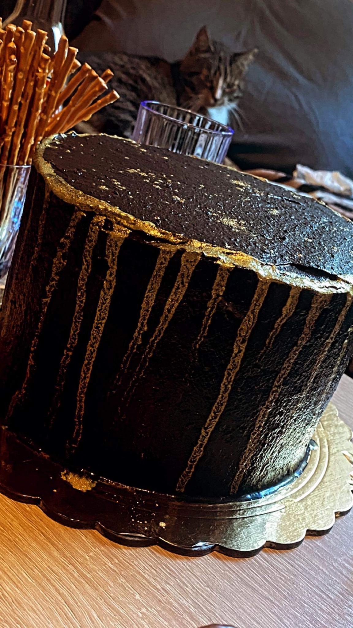 Ciemny tort z abstrakcyjnymi złotymi zdobieniami, na złotym podkładzie, w tle szklanka ze słonymi paluszkami, szklanka i kot.