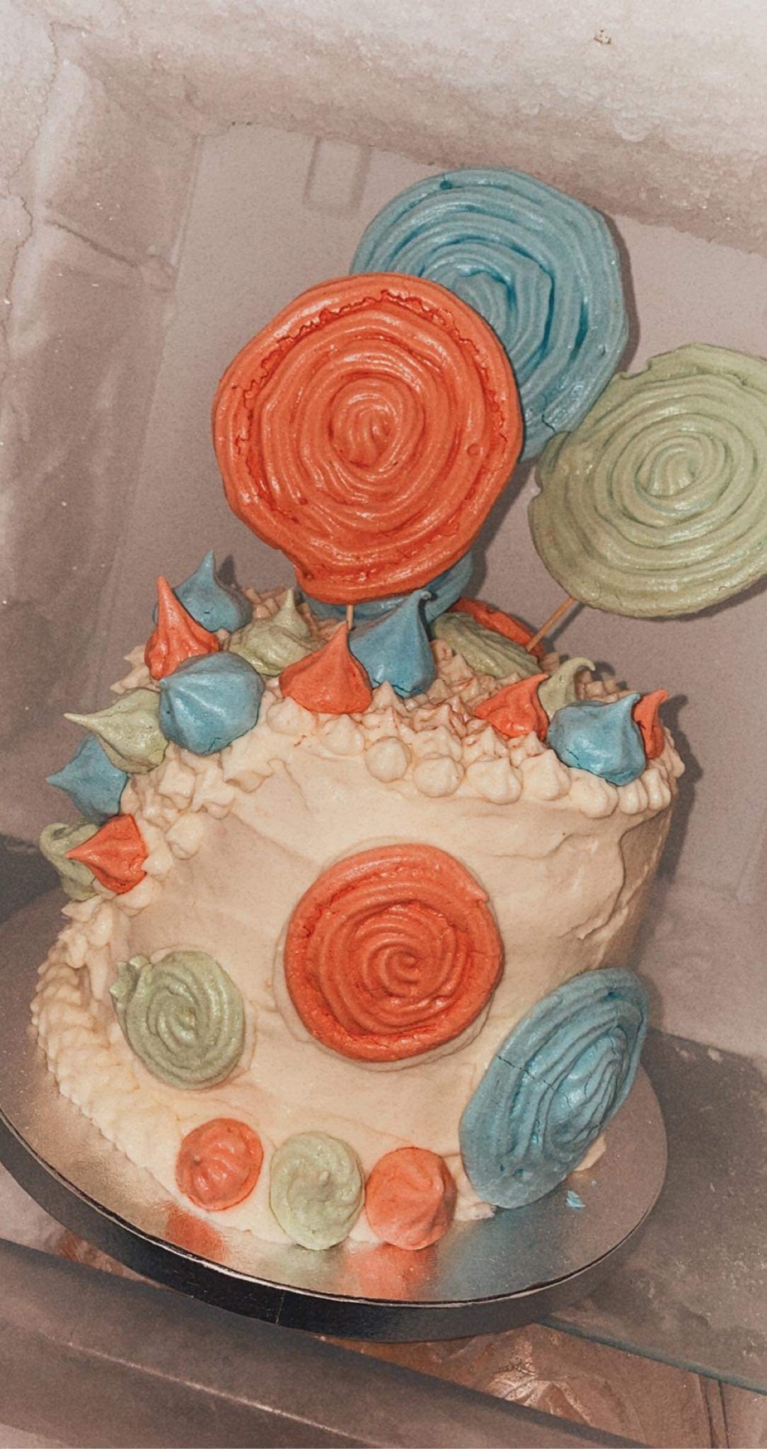 Tort z kremem i kolorowymi bezami w kształcie lizaków, umieszczony na srebrnej paterze, sfotografowany w chłodnym otoczeniu.