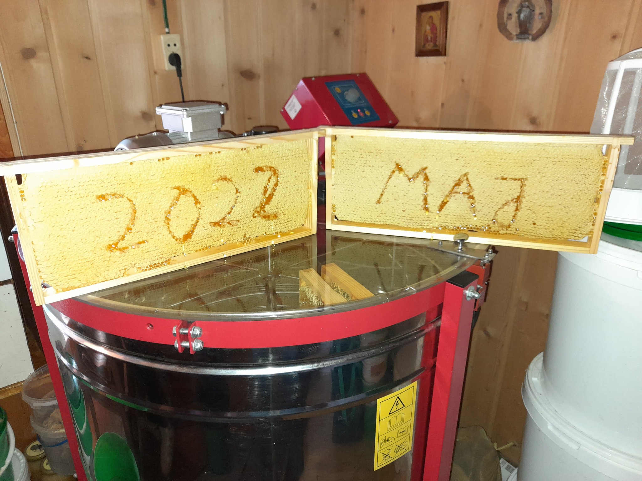 Dwa ramki z miodem na miodarce, na jednej napis '2022' uformowany z miodu, na drugiej 'MAJ', w tle drewniana boazeria i sprzęt pszczelarski.
