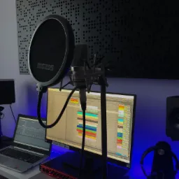 Mikrofon studyjny z filtrem pop na tle monitora z oprogramowaniem DAW, głośniki studyjne i laptop z interfejsem audio na biurku oświetlonym niebieskim światłem.