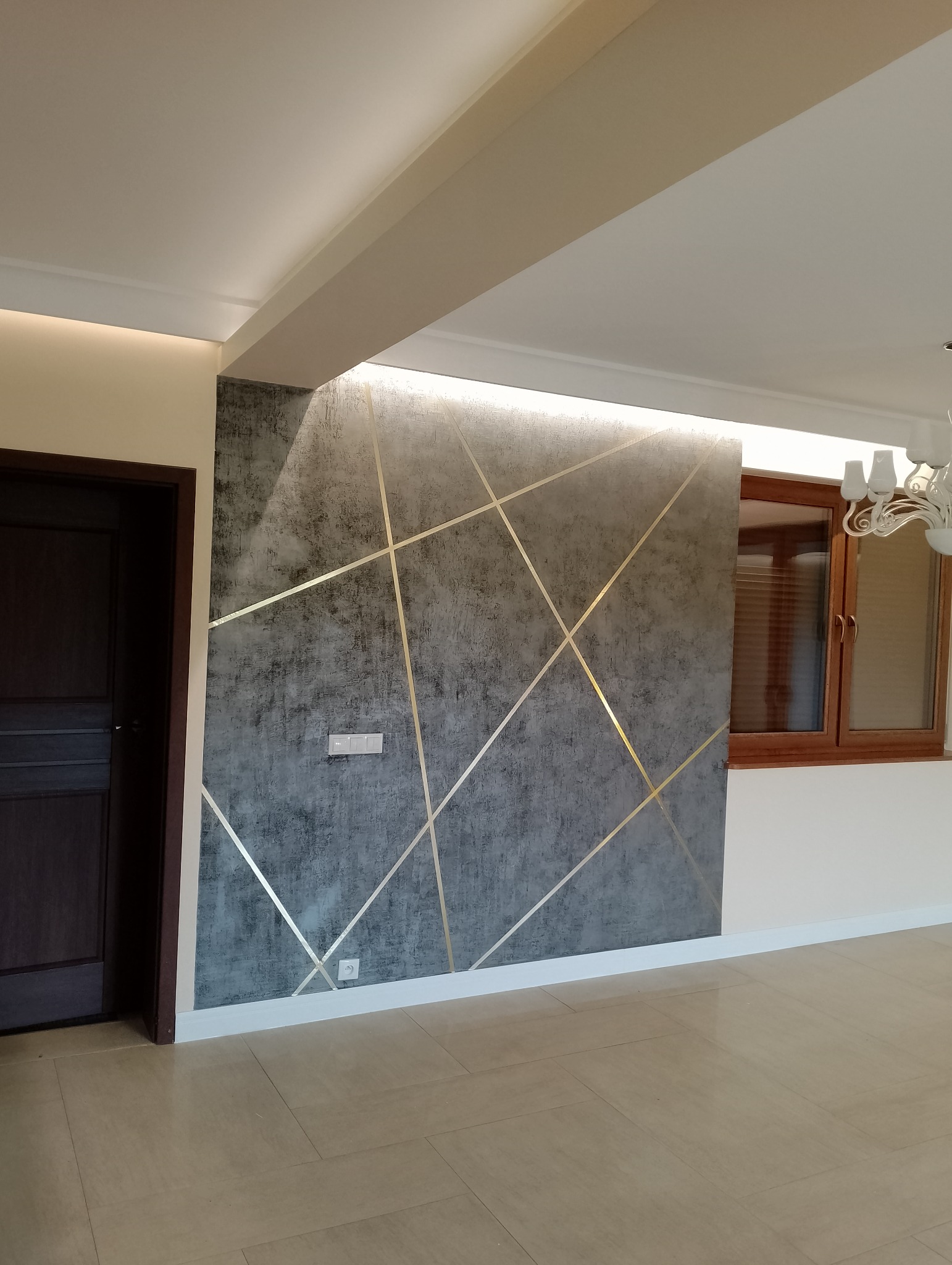 Szara ściana z dekoracyjnymi, złotymi liniami w nowoczesnym wnętrzu, widoczny fragment sufitu z oświetleniem LED, brązowe drzwi i beżowa podłoga z płytek.