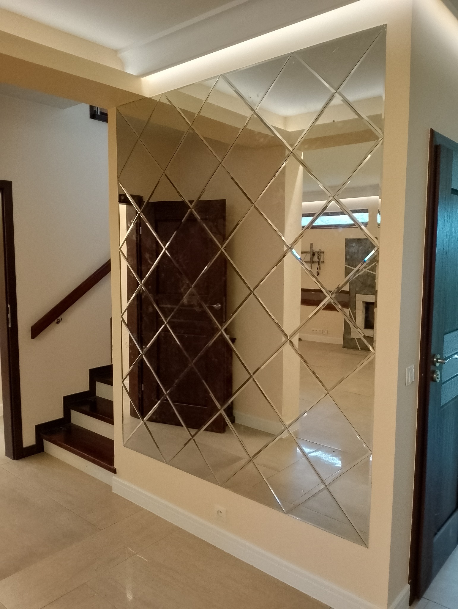 Ściana wyłożona dekoracyjnymi lustrami w kształcie rombów, odbijająca fragment schodów i sąsiedniego pomieszczenia z kominkiem.