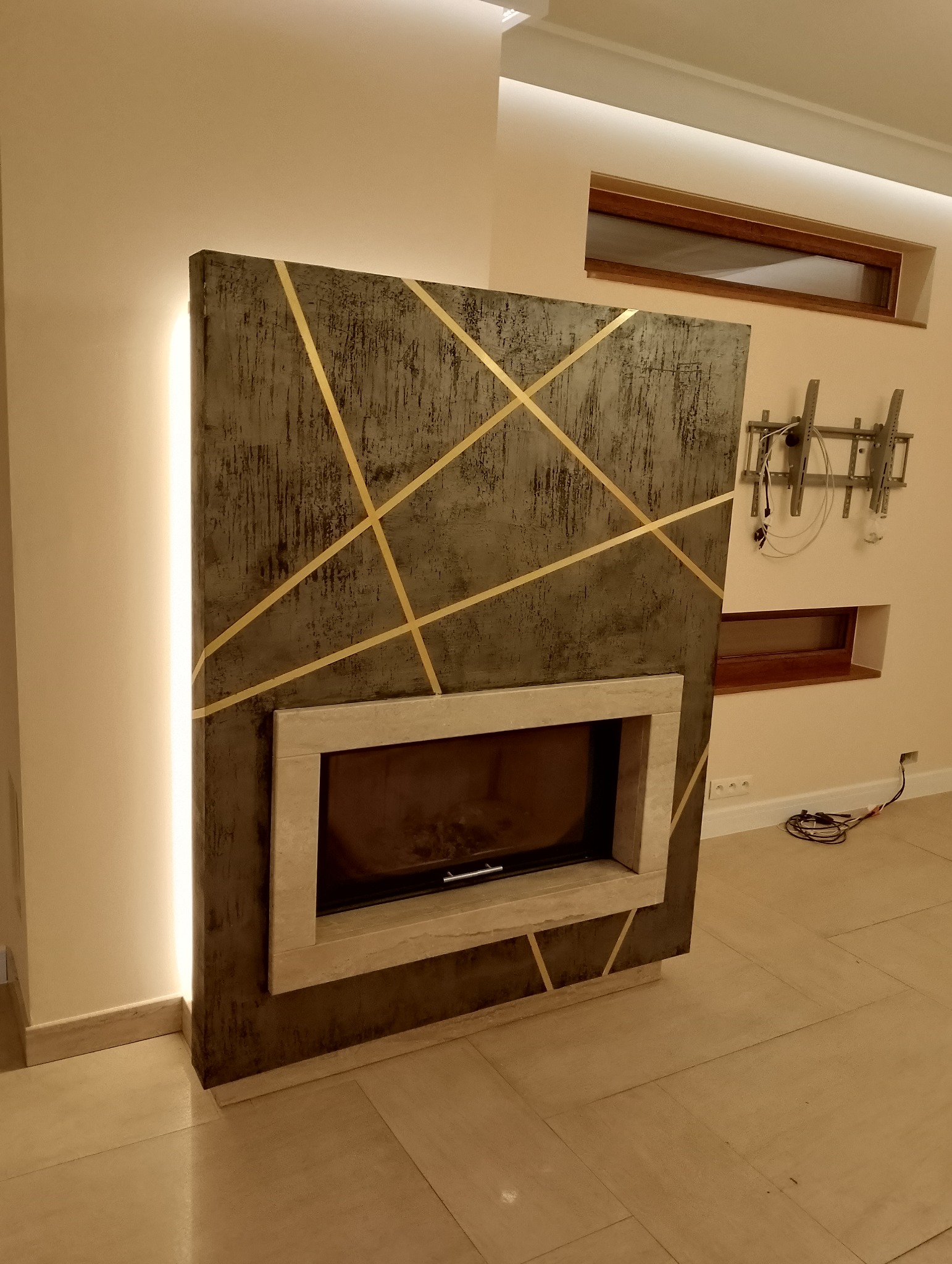 Obudowa kominka z szarego betonu architektonicznego z geometrycznymi, złotymi wstawkami, podświetlana listwą LED, w nowoczesnym salonie.