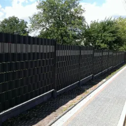 Długi, czarny płot panelowy z szarymi akcentami, oddzielający posesję od chodnika i ulicy w słoneczny dzień.