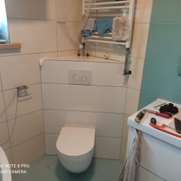Krzysztof Zarzyczny Usługi remontowo budowlane - Biała wisząca toaleta ze spłuczką podtynkową na tle białych płytek, na umywalce narzędzia i akcesoria łazienkowe, w tle grzejnik drabinkowy i lustro.