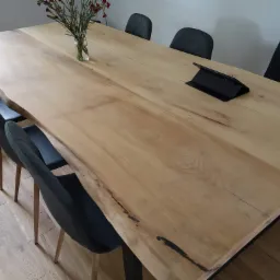 Surowy stół z jasnego drewna z naturalnymi krawędziami, czarne metalowe nogi, otoczony szarymi krzesłami, na blacie wazon z kwiatami i tablet.