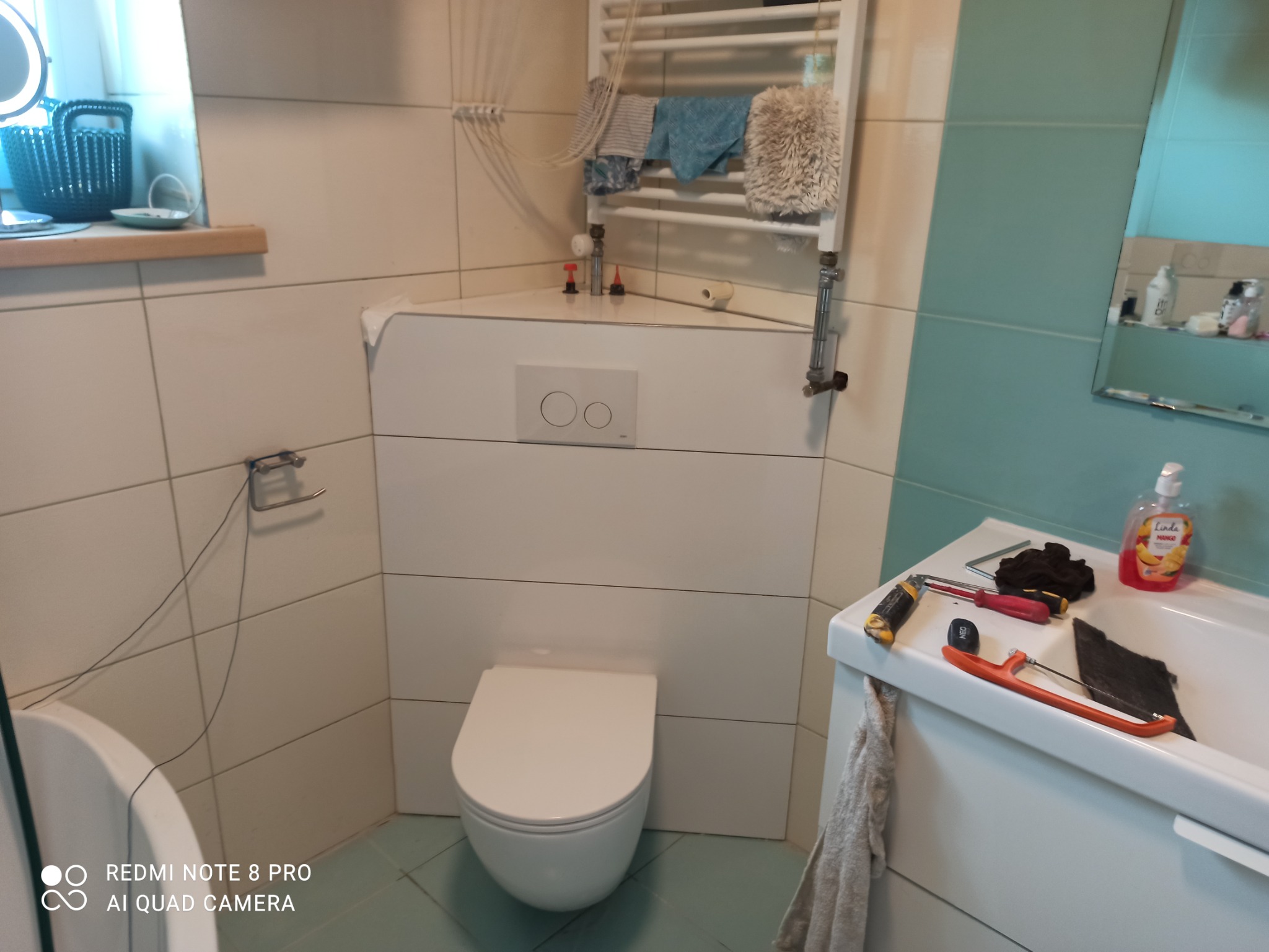 Biała wisząca toaleta ze spłuczką podtynkową na tle białych płytek, na umywalce narzędzia i akcesoria łazienkowe, w tle grzejnik drabinkowy i lustro.