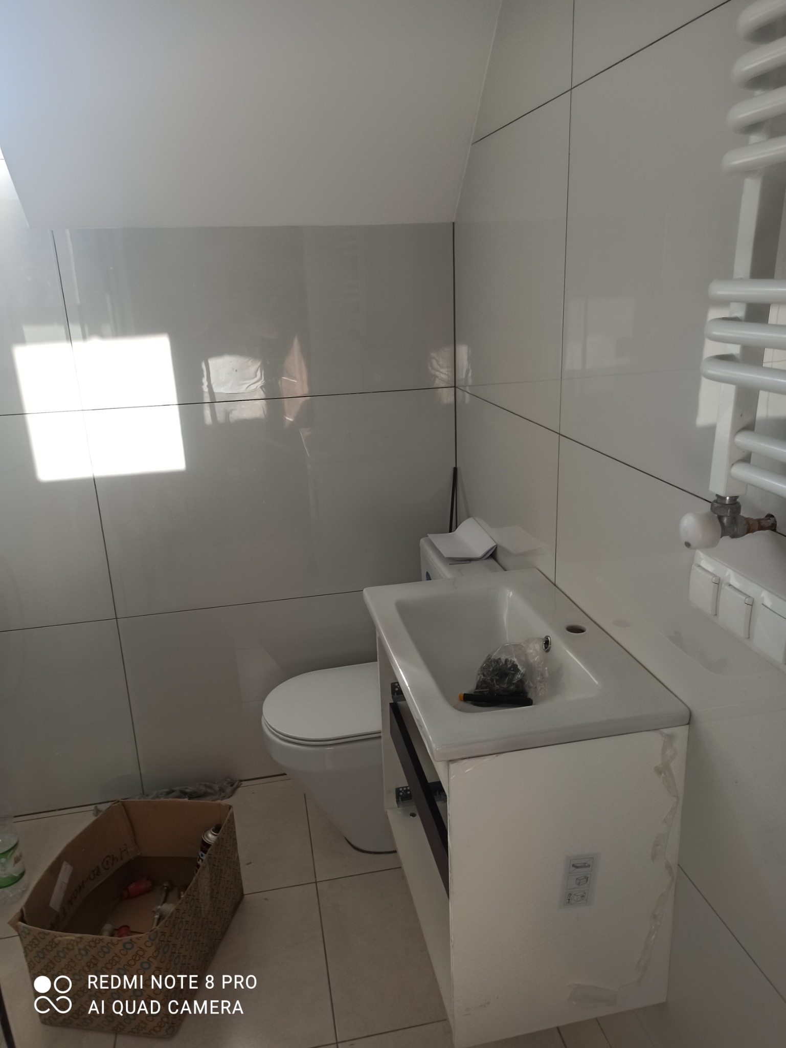 Biała łazienka w trakcie wykańczania, widoczna umywalka z szafką, toaleta, grzejnik drabinkowy i karton z narzędziami na podłodze. Na umywalce leży woreczek z wkrętami i śrubokręt.