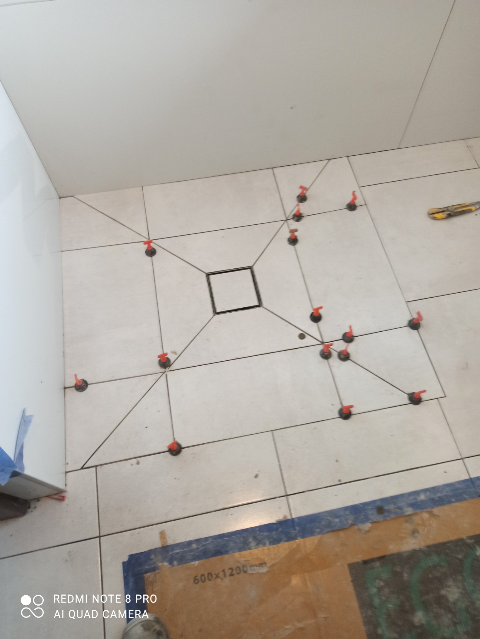 Białe płytki podłogowe 600x1200 mm w trakcie układania w łazience z centralnym odpływem liniowym, widoczne czerwone klipsy systemu poziomowania płytek oraz nóż do cięcia płytek.