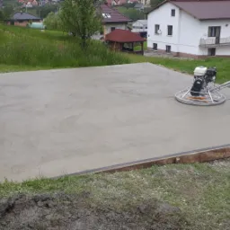 Wyrównywanie wylewki jastrychowej w Iwkowej za pomocą zacieraczki mechanicznej na tle zieleni i zabudowań.