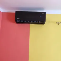 Czarny klimatyzator marki Rotenso na ścianie pomalowanej w kolorze żółtym i czerwonym, wyświetlający temperaturę 23 stopnie.