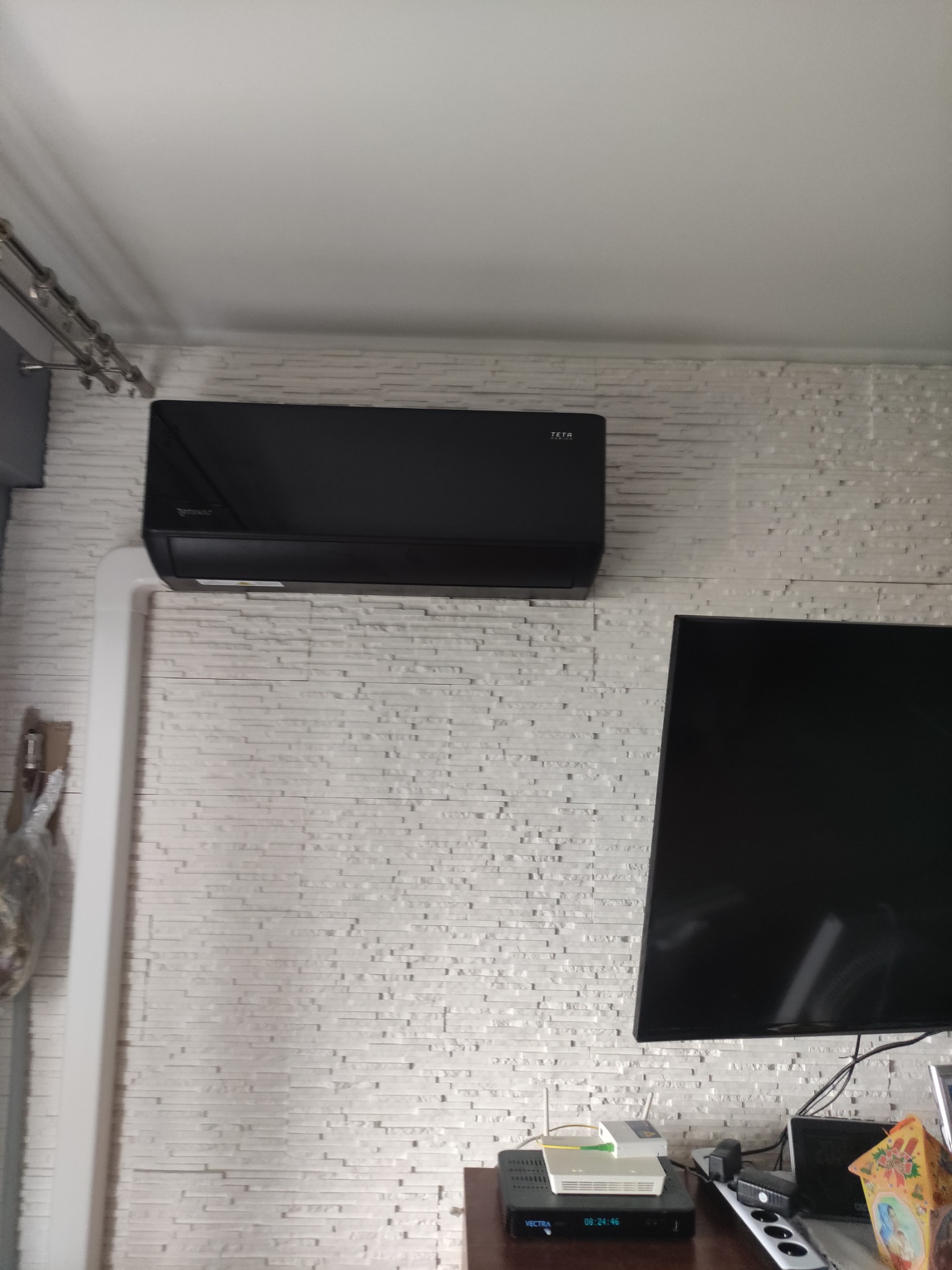 Czarny klimatyzator TETA zamontowany na ścianie z białej cegły dekoracyjnej, widoczny fragment telewizora i dekoder Vectra