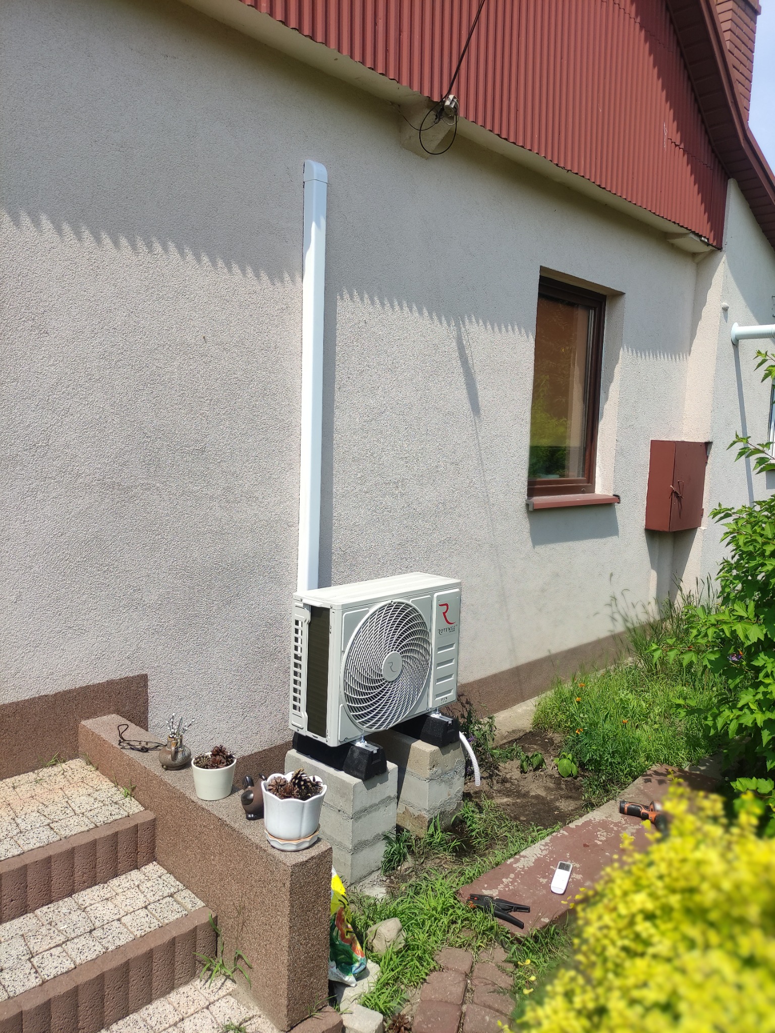 Zewnętrzna jednostka klimatyzacji zamontowana na betonowych bloczkach przy ścianie domu, z widoczną rurą odprowadzającą skropliny i elementami instalacji. Obok schody z dekoracjami i narzędzia...